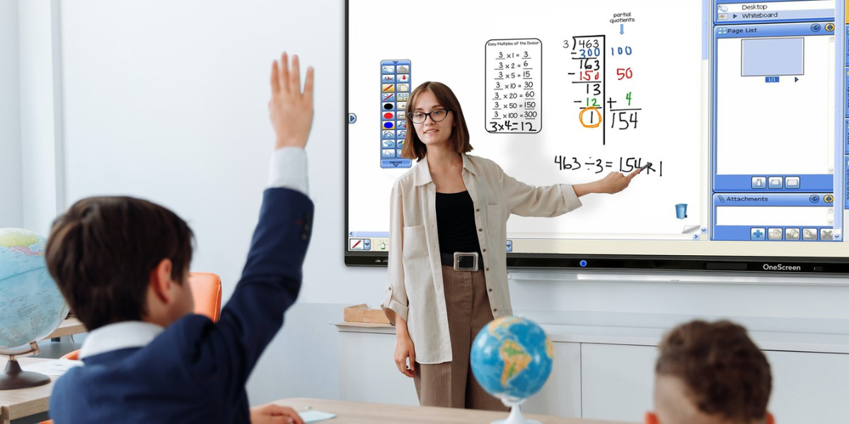 Teacher points to equation on OneScreen interactive whiteboard as young student raises hand.