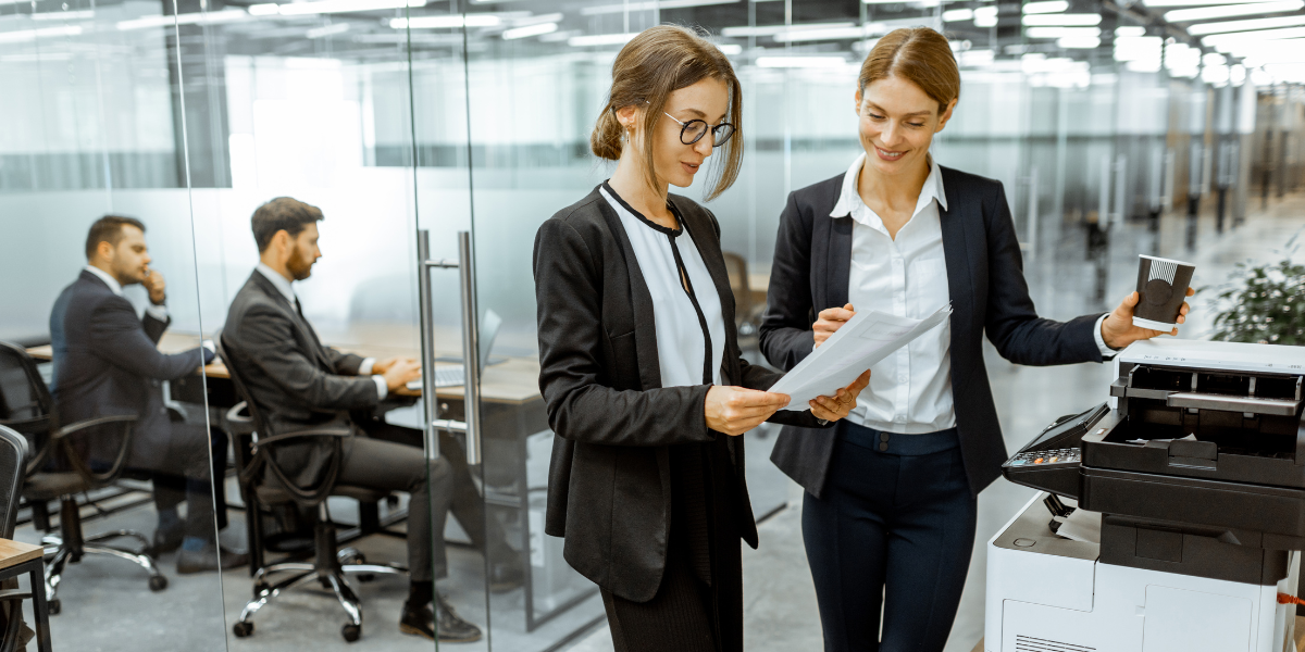 Two women using printer in office while meeting happens in background