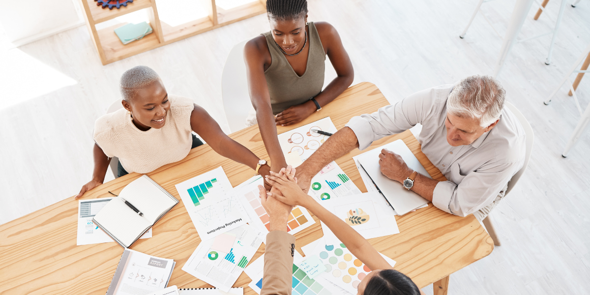 Team joining hands across meeting table with report papers spread over it