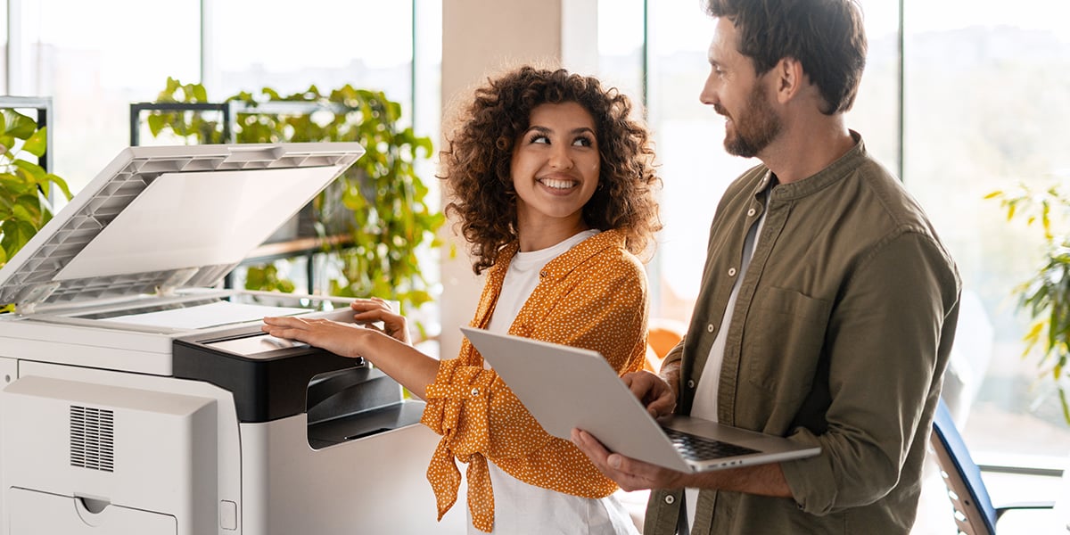 Two business partners smiling and collaborating while using a printer and laptop